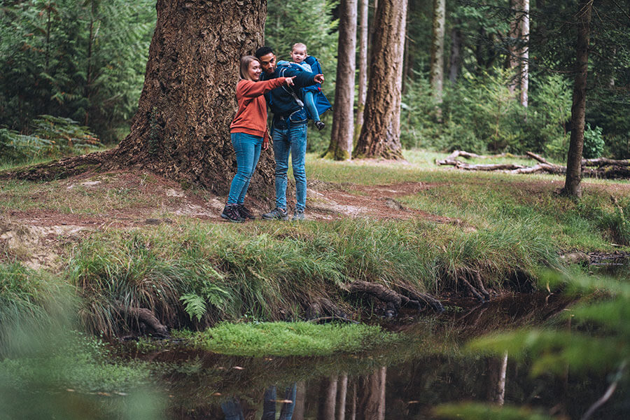 Familie mit Kind in Kindertrage an einem Bachlauf im Wald unterwegs