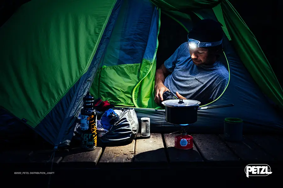 Mann mit Stirnlampe auf dem Kopf aus Zelt heraus auf Kochstelle schauend